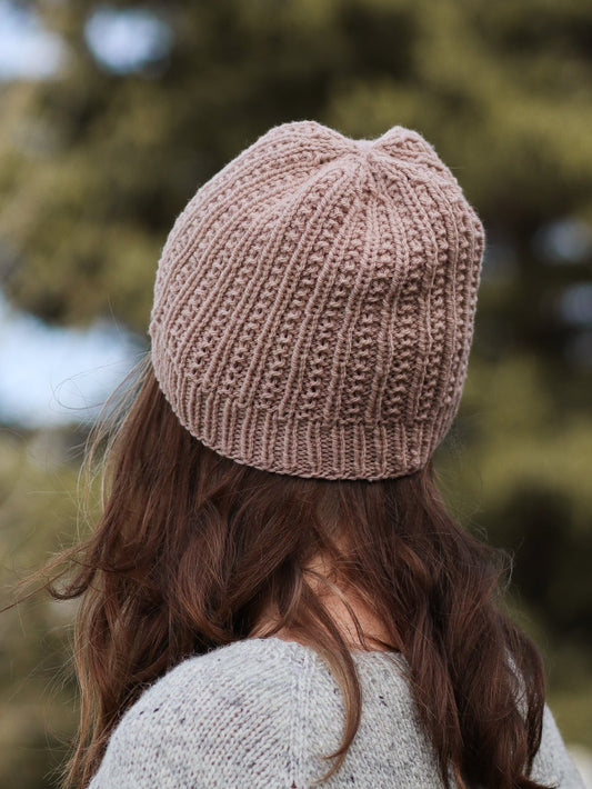 Person wearing a knitted pink beanie and gray sweater with blurred trees in the background